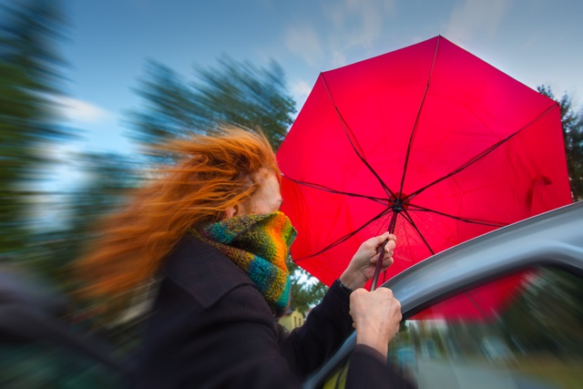 Sturm. Frau steigt aus Auto, roter Regenschirm fliegt fast weg
